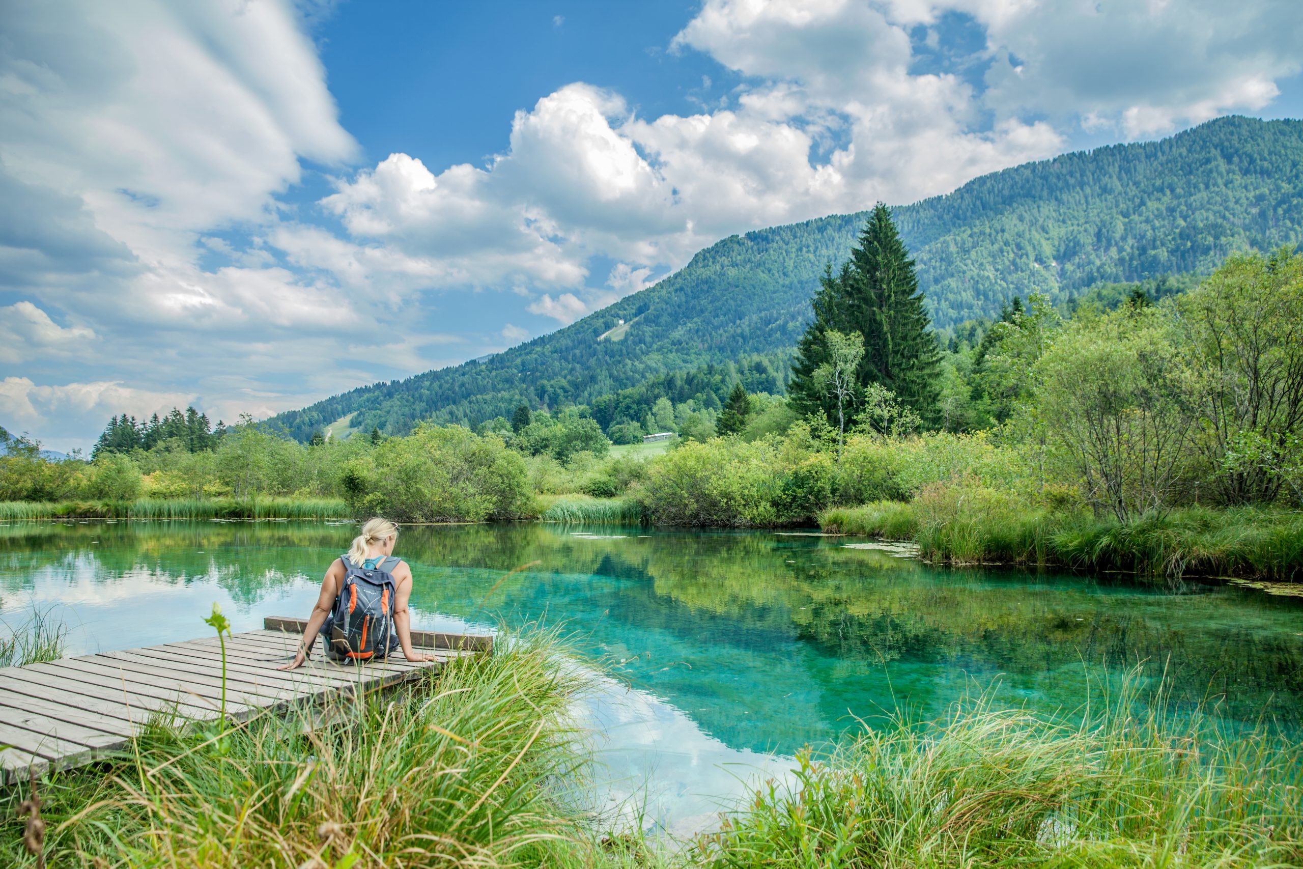 A picture of a female sitting on a wooden bridge against an emerald lake with a breathtaking nature in Slovenia